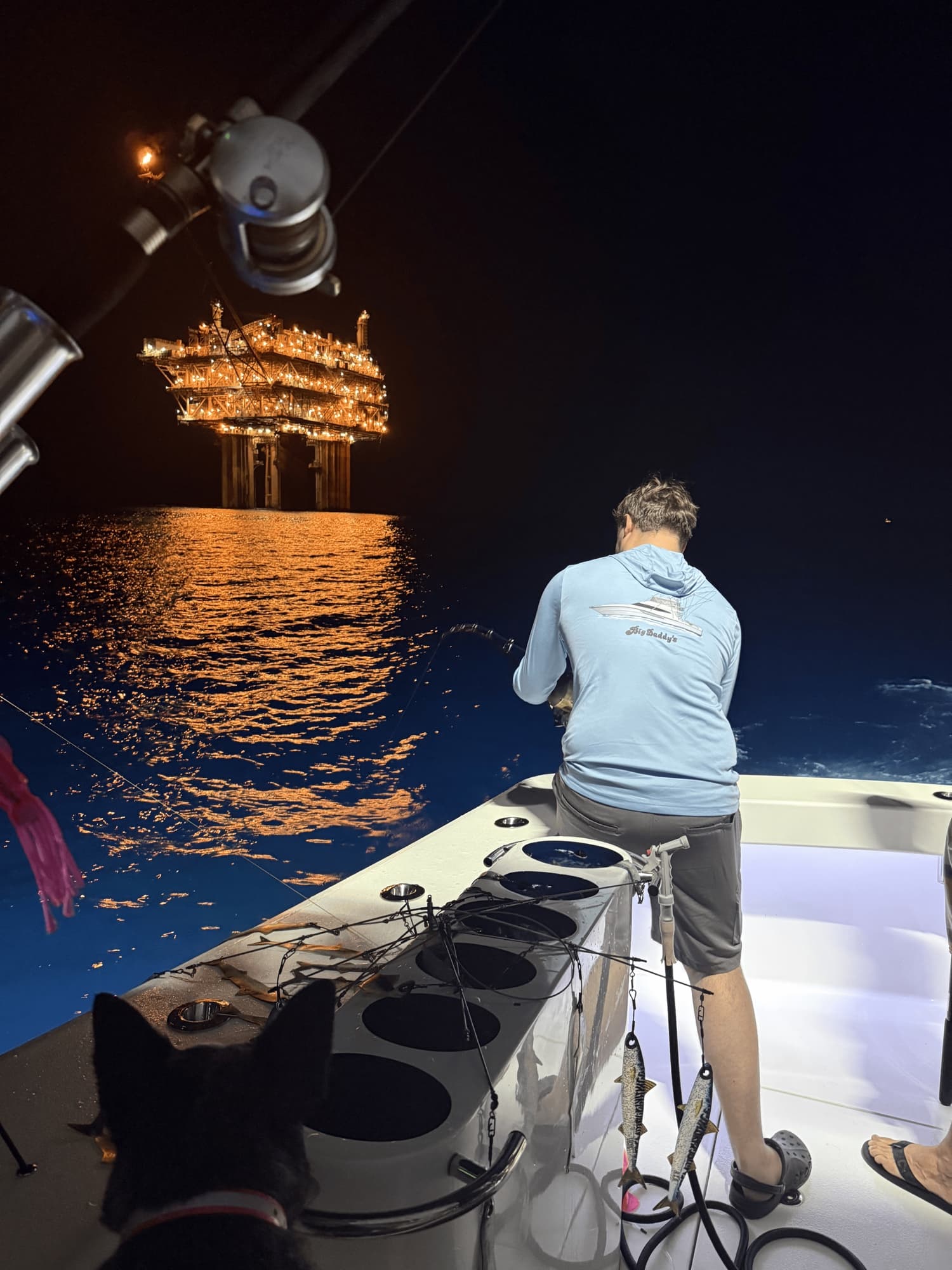 Night fishing under a lit Gulf oil platform — angler at the rod, the boat dog watching from the cockpit